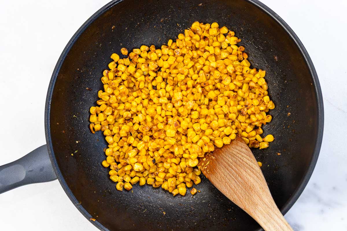 Overhead view of sauteed corn in a small wok style pan with a wooden spoon.