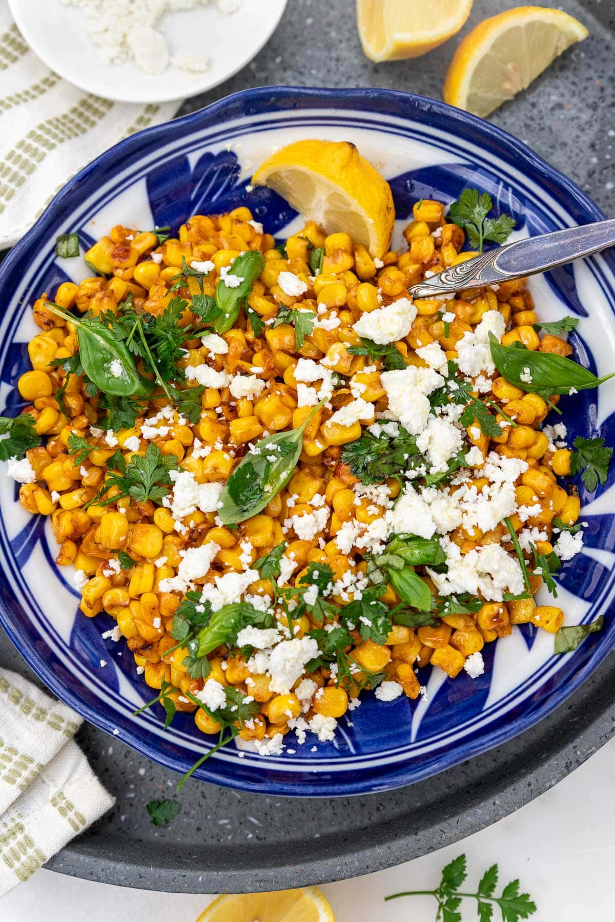 An overhead view of a blue dish with sauteed corn, feta cheese and herbs, on a grey tray and with a cream and green tea towel.