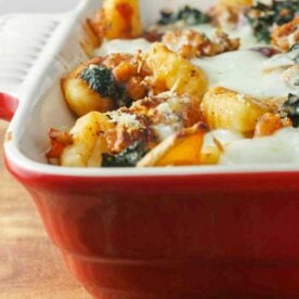 Closeup of a red baking dish of butternut squash gnocchi with sausage and kale on a wooden board.