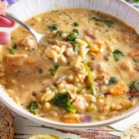 A white bowl of creamy chicken orzo soup with someone scooping up a spoonful, and with ingredients scattered around it.