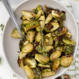 The roasted brussels in a bowl with a spoon, with fresh thyme on top and on a white and green tea towel.