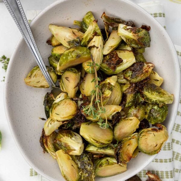 The roasted brussels in a bowl with a spoon, with fresh thyme on top and on a white and green tea towel.