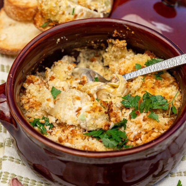 Creamy baked artichoke gratin with golden breadcrumbs on top with a spoon in it and in a small brown ceramic baking dish.