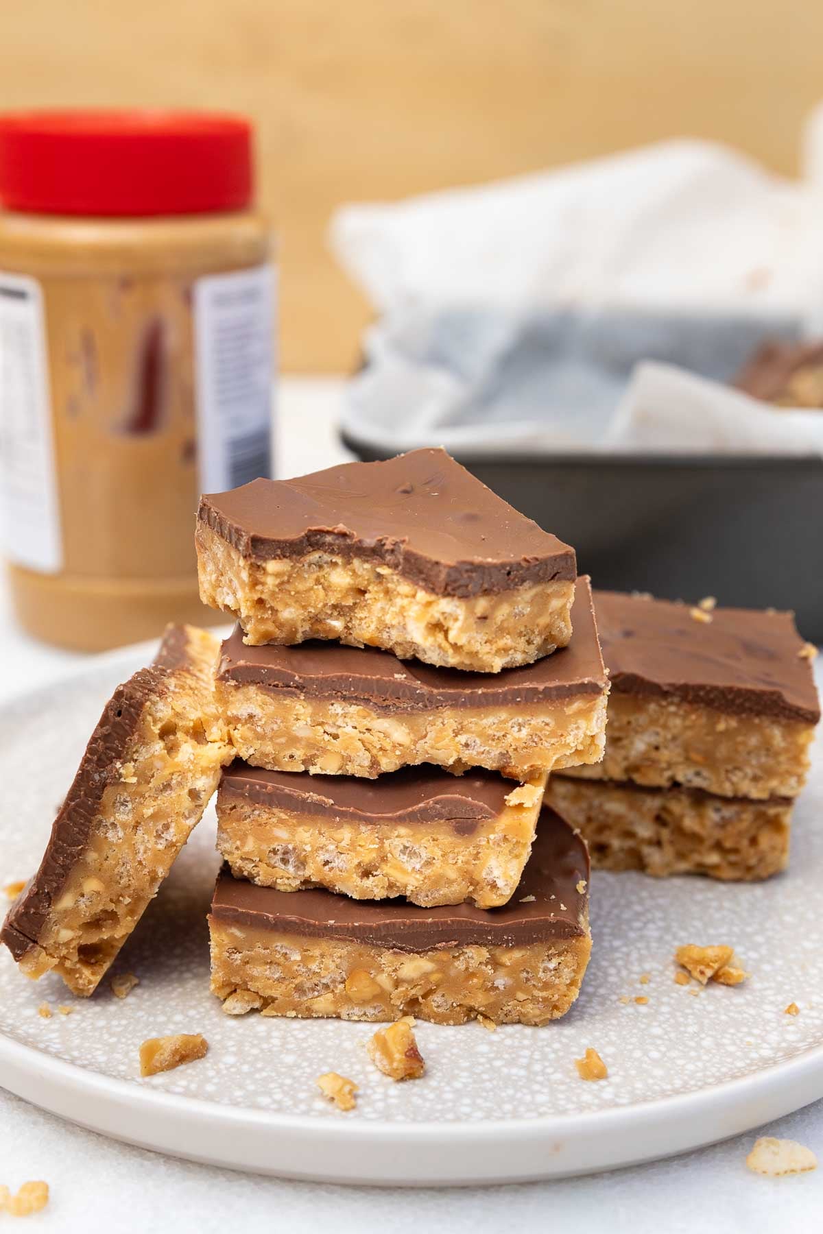 A stack of the crunch bars on a plate with more at the sides and behind, with a jar of peanut butter behind.
