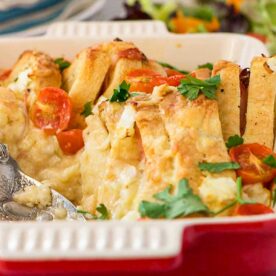 Closeup of a red baking dish with a ham, cheese and egg bake in it and a serving spoon, with a simple salad and tea towel in the background.