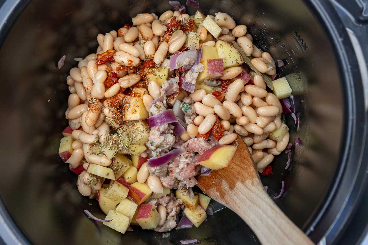 Browned Italian sausage, white beans, herbs, potatoes, onion and herbs in a crockpot with a wooden spoon.
