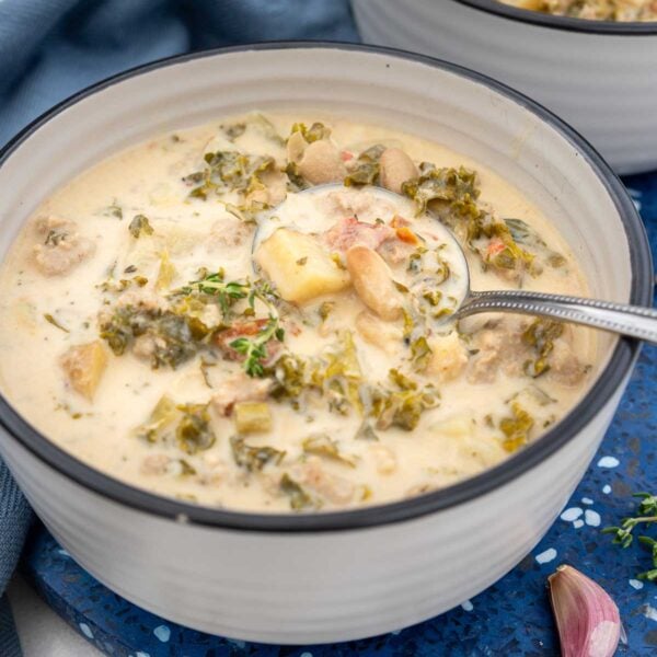 A bowl of crock pot zuppa toscana on a blue background with blue tea towel and with a metal spoon in it.