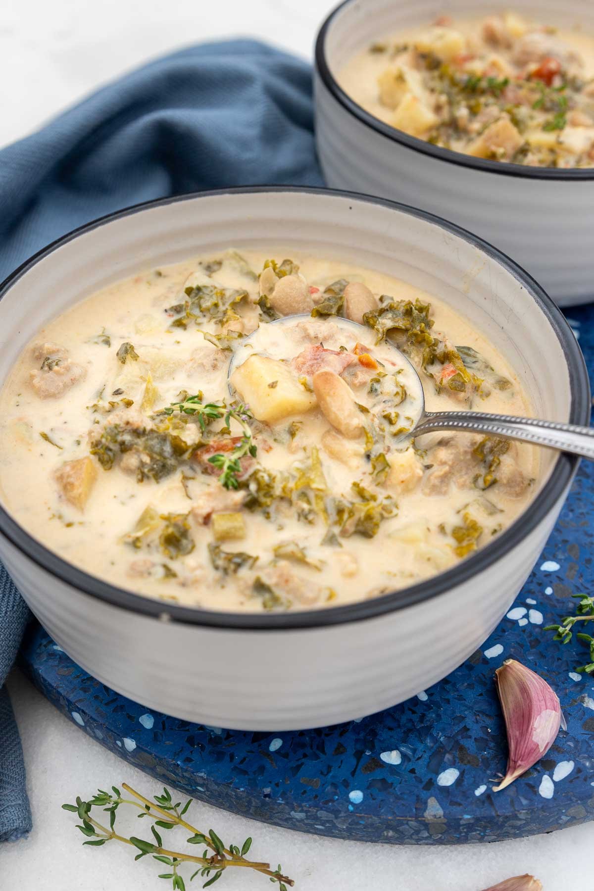 A bowl of crock pot zuppa toscana on a blue background with blue tea towel and with a metal spoon in it.