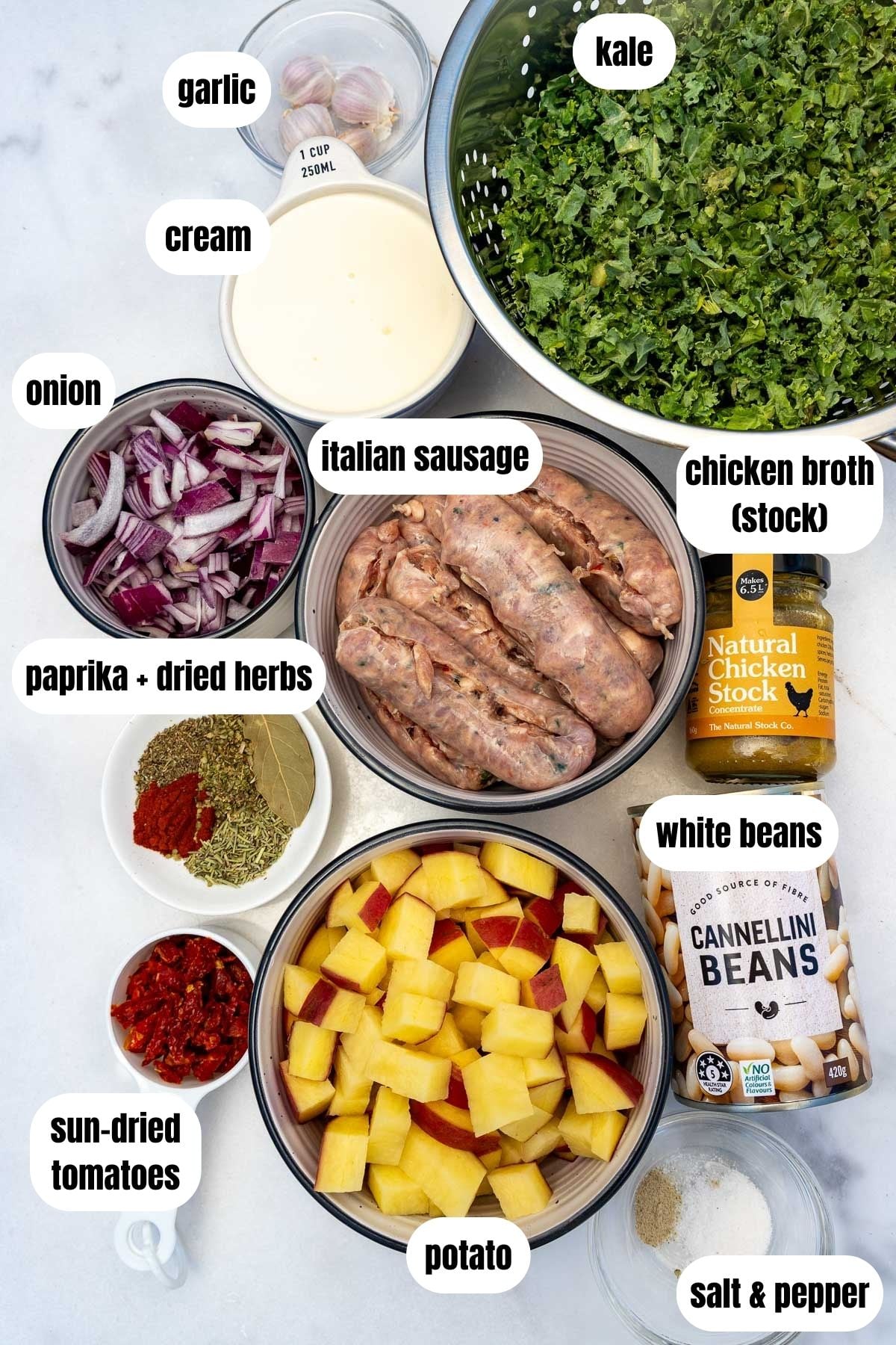 All ingredients needed to make crock pot zuppa toscana labelled including Italian sausages, diced potatoes, sun-dried tomatoes, salt and pepper, white beans, paprika and dried herbs, chicken broth, onion, garlic, kale and garlic.