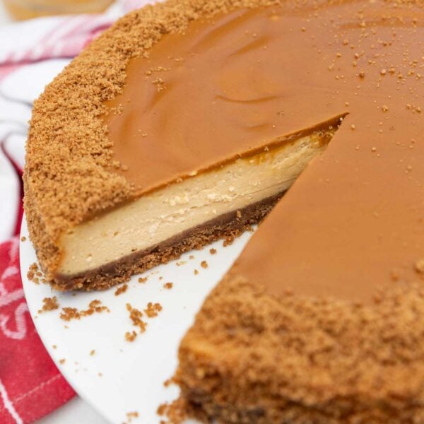 Closeup of part of a whole Biscoff cheesecake with a piece cut out of it so you can see the inside, on a white cake stand with a read tea towel.
