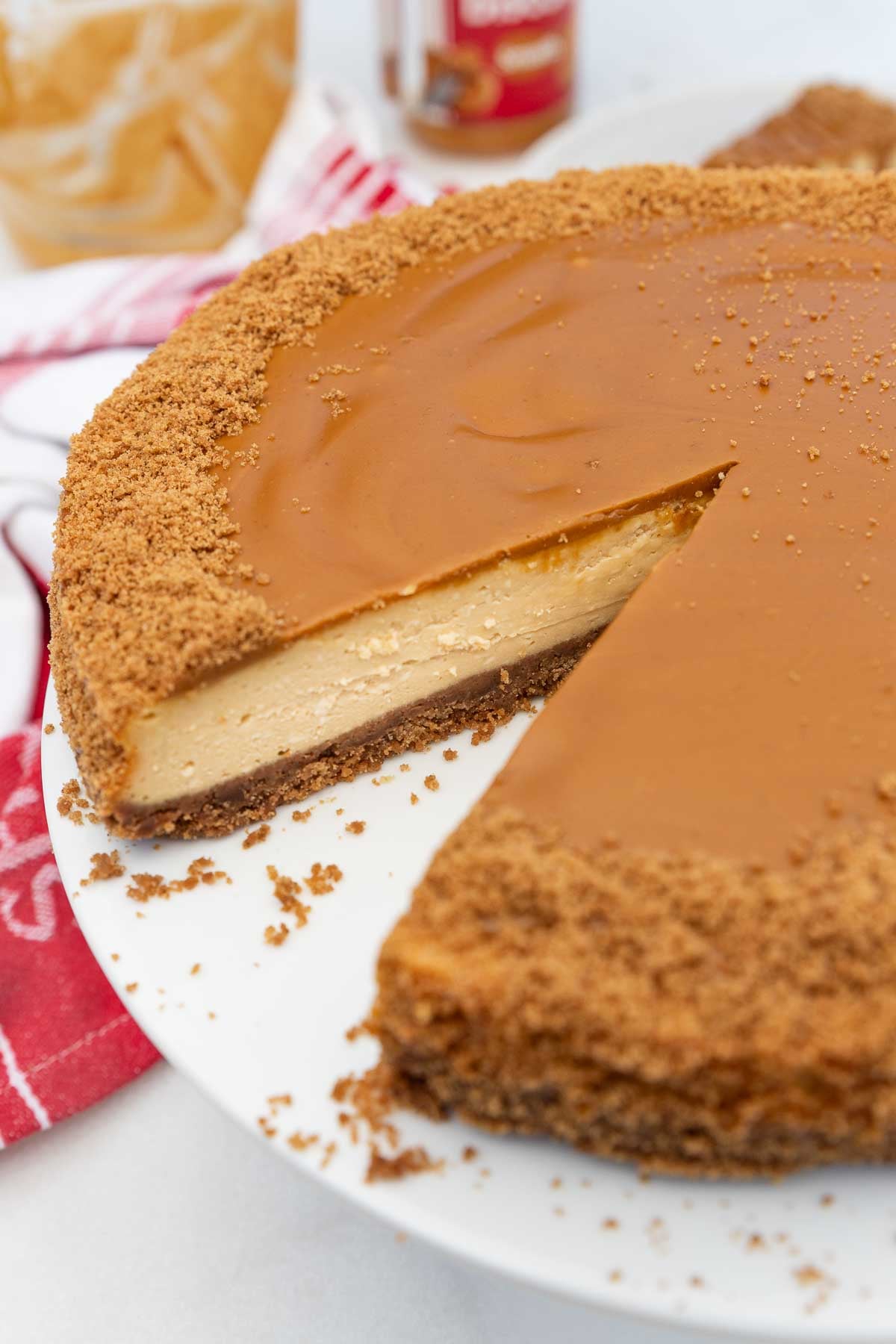 Closeup of part of a whole Biscoff cheesecake with a piece cut out of it so you can see the inside, on a white cake stand with a read tea towel.