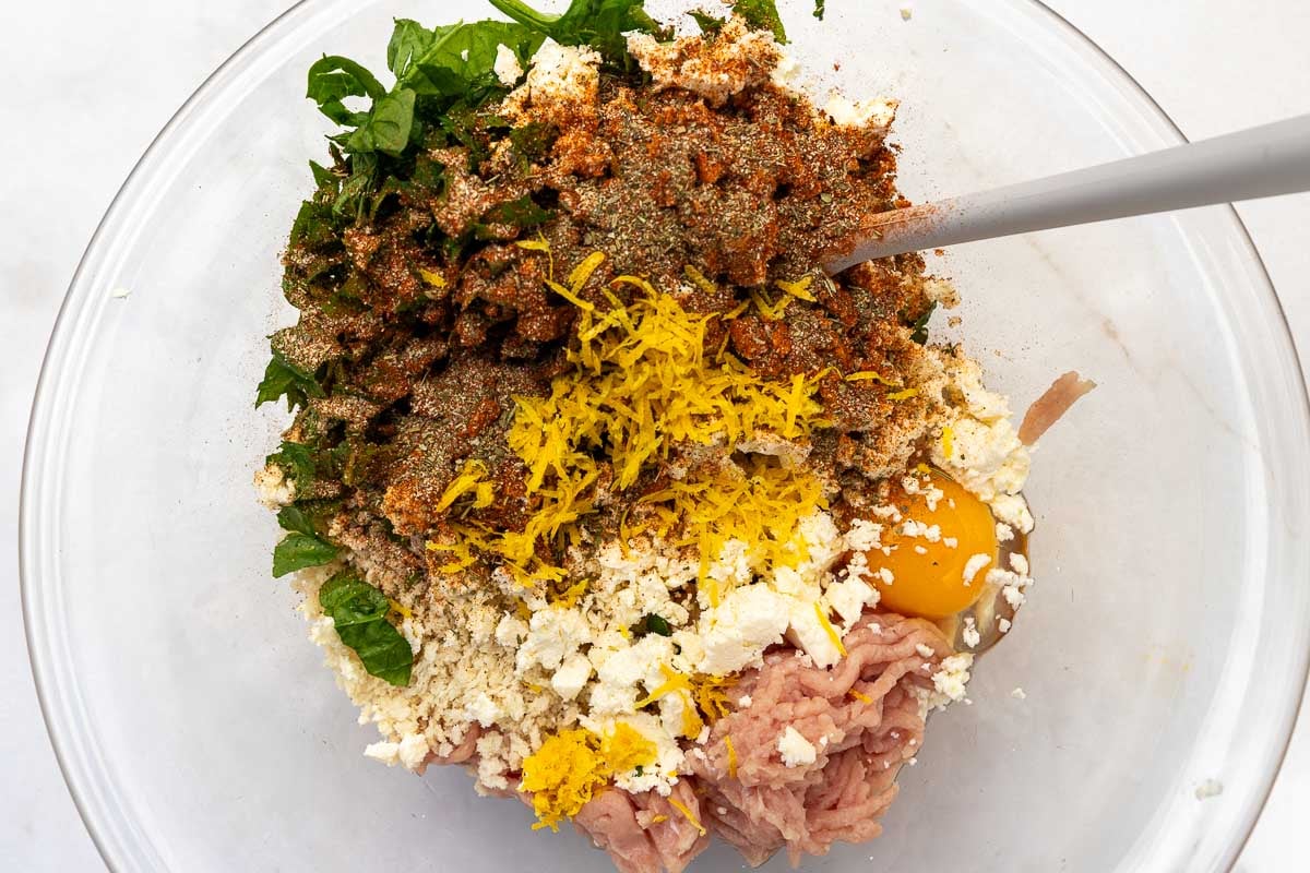All the ingredients needed to make the chicken burgers in a clear bowl with a spoon, ready to be mixed.