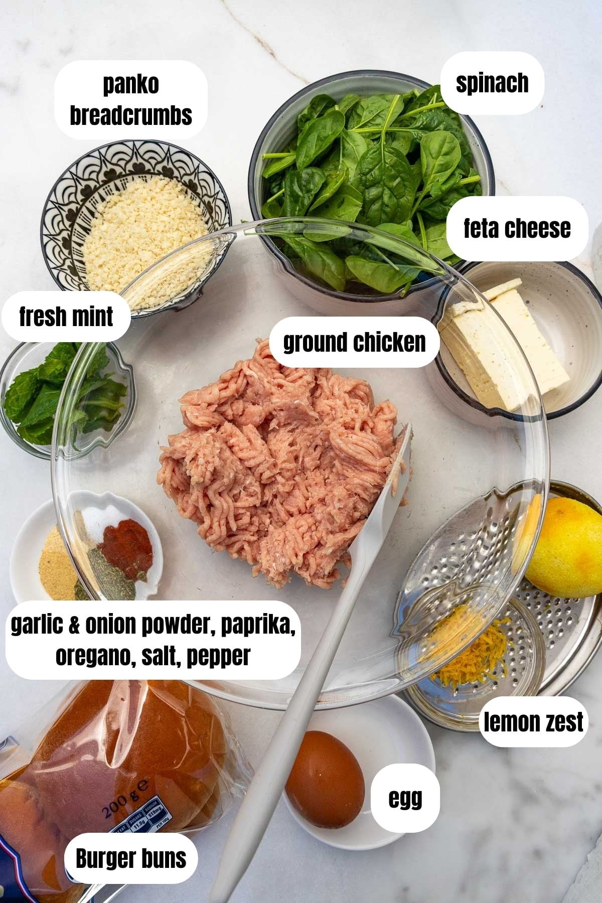 All the ingredients needed to make the burgers labelled, including ground chicken, burger buns, an egg, lemon zest, seasoning, fresh mint, feta cheese, baby spinach, panko breadcrumbs and fresh mint.