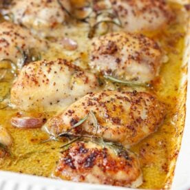 A closeup of honey mustard chicken thighs just baked in a red baking dish, with fresh rosemary and garlic visible on top.