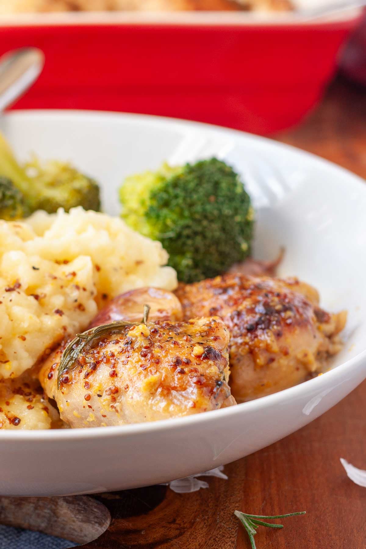 Part of a white bowl with two whole skinless chicken thighs with honey mustard sauce, with mashed potatoes and broccoli.