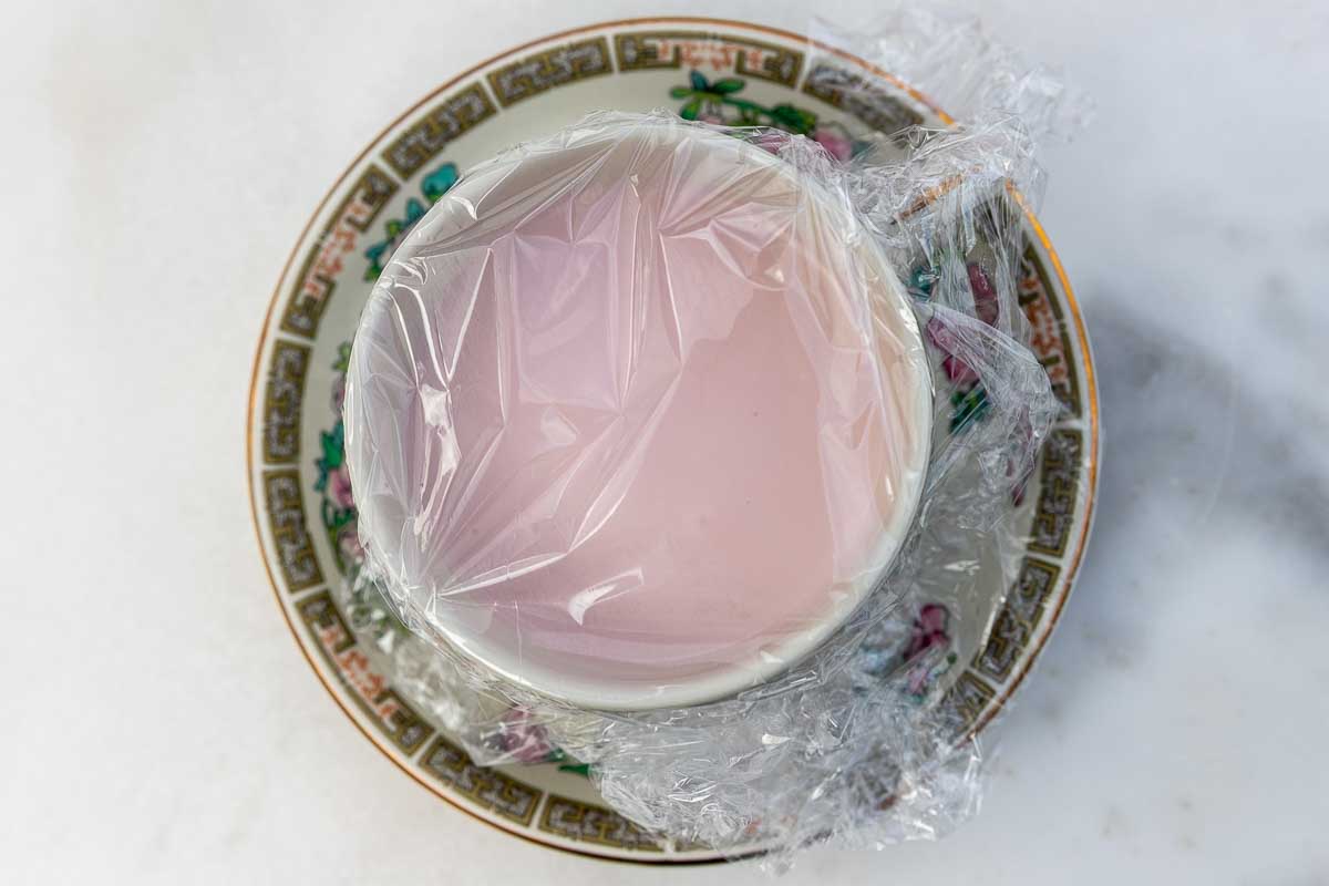 An overhead of the dessert in a pretty teacup on a saucer, with plastic wrap over the top.