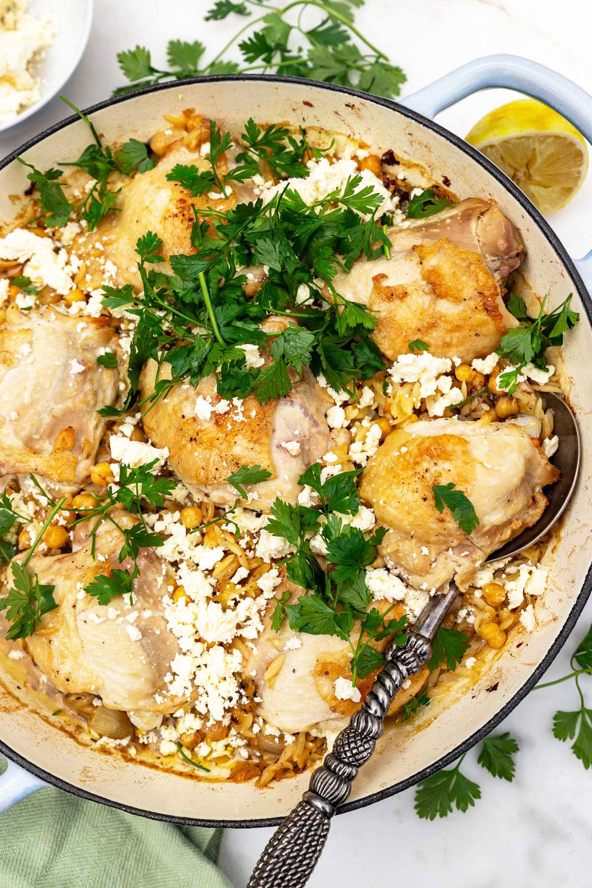 Overhead of a whole pan of chicken and orzo with fresh parsley and feta on top, and a decorative spoon in it.