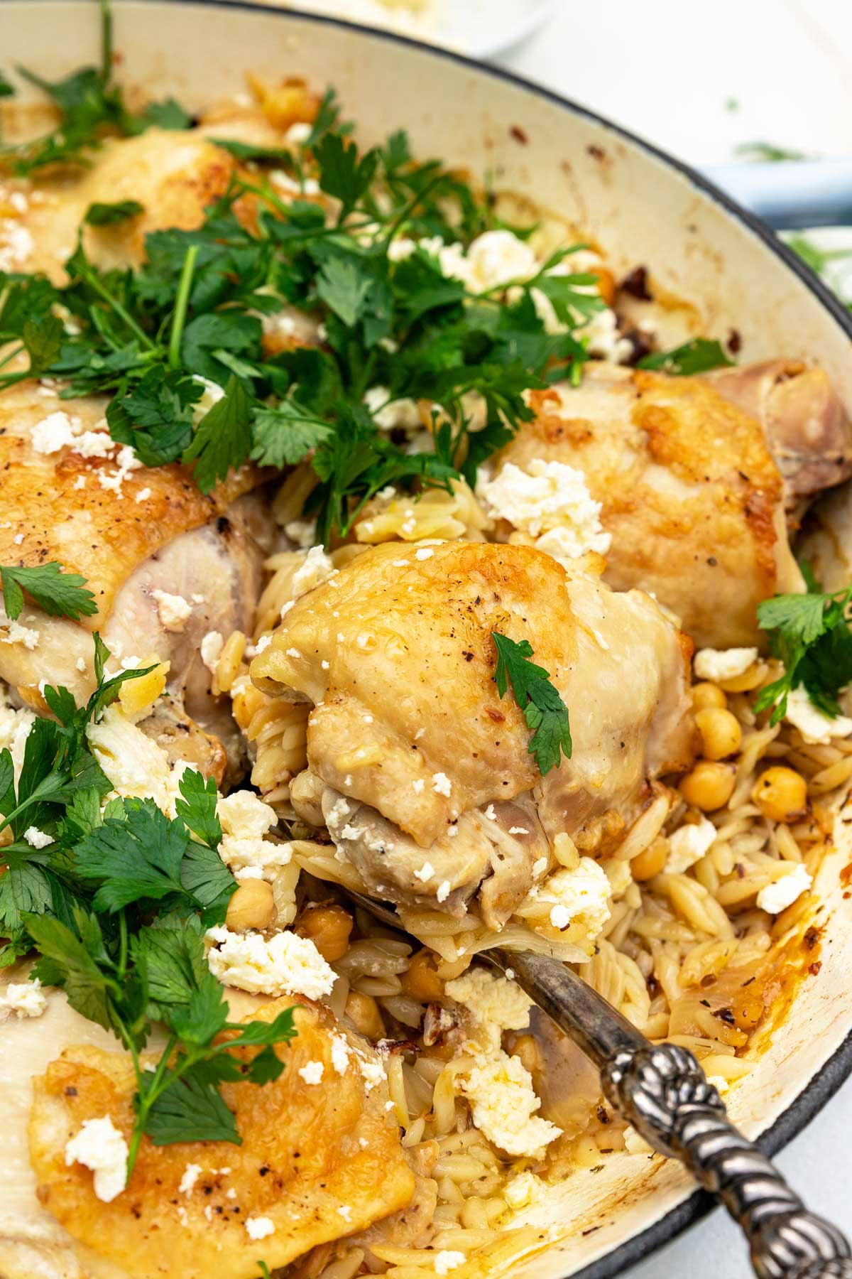 Closeup of a decorative spoon in the pan scooping out a chicken thigh with the orzo, chickpeas and toppings around it.