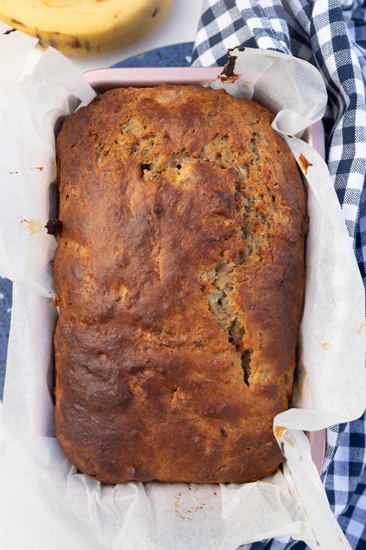 The baked banana loaf still in the baking pan, with a blue checked tea towel at the side.