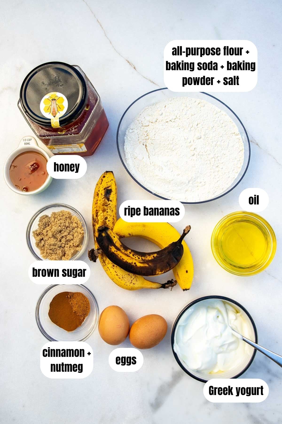 All the ingredients to make Greek yogurt banana bread, labelled, including a flour mixture, eggs, Greek yogurt, oil, cinnamon and nutmeg, ripe bananas, brown sugar, and honey.