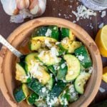 A small earthenware dish with sauteed zucchini in it, sprinkled with parmesan and with a metal spoon in the dish and ingredients around it.