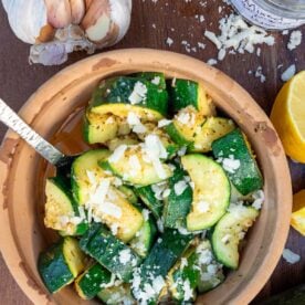 A small earthenware dish with sauteed zucchini in it, sprinkled with parmesan and with a metal spoon in the dish and ingredients around it.