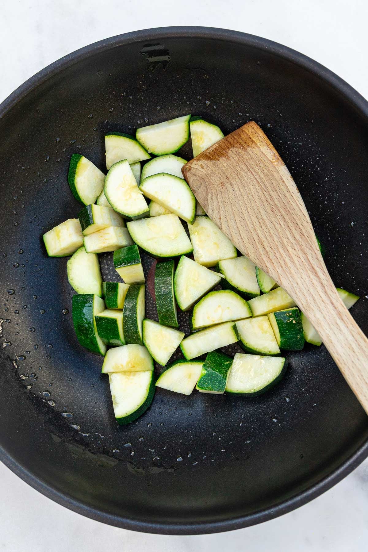 Overhead view of quartered slices of zucchini in a pan with olive oil and a wooden spatula.