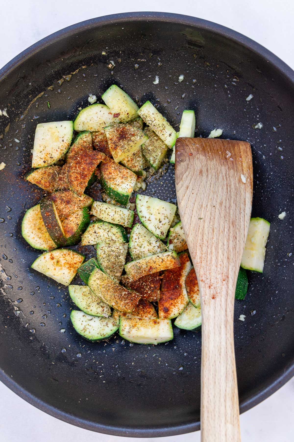 Slices of raw zucchini, quartered, in a saute pan with dried herbs and spices, with a wooden spatula.