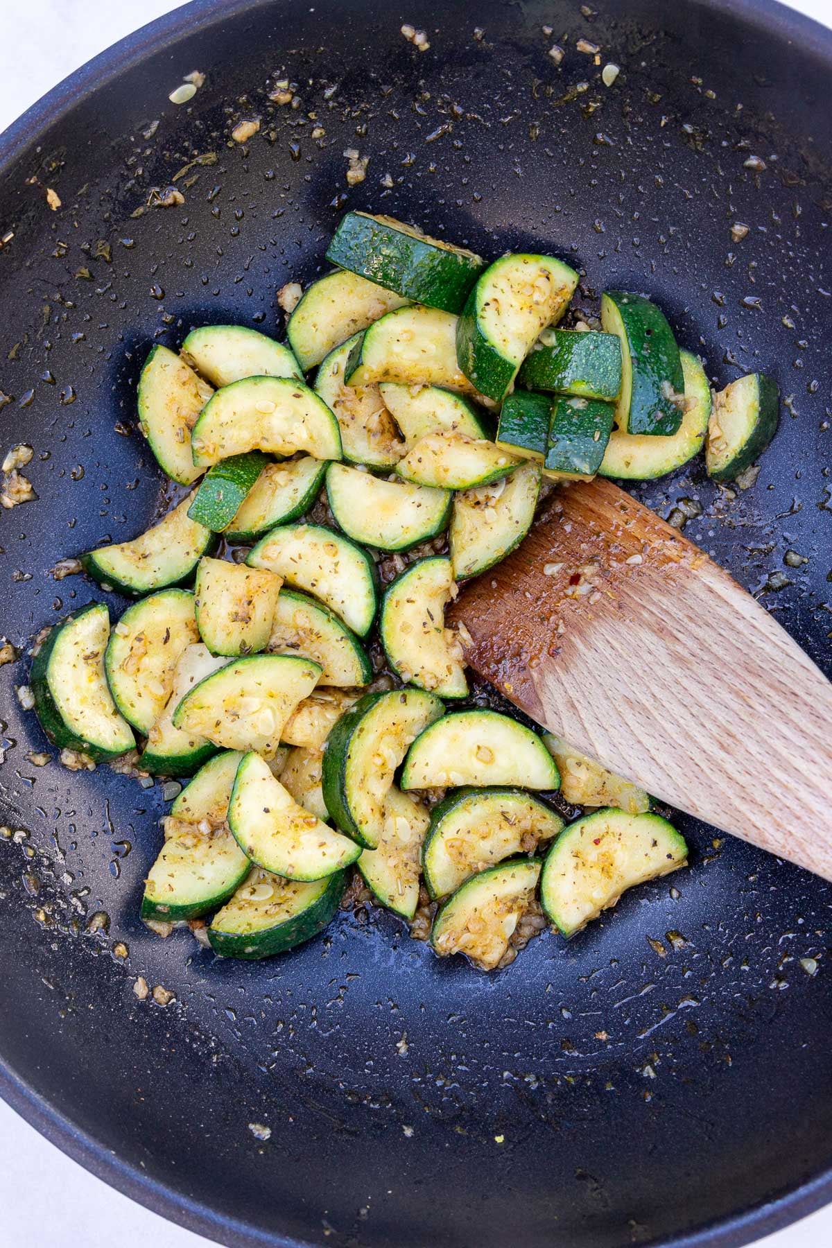 Overhead view of zucchini sauteed in a pan with garlic, herbs and other simple flavors, plus a wooden spatula.