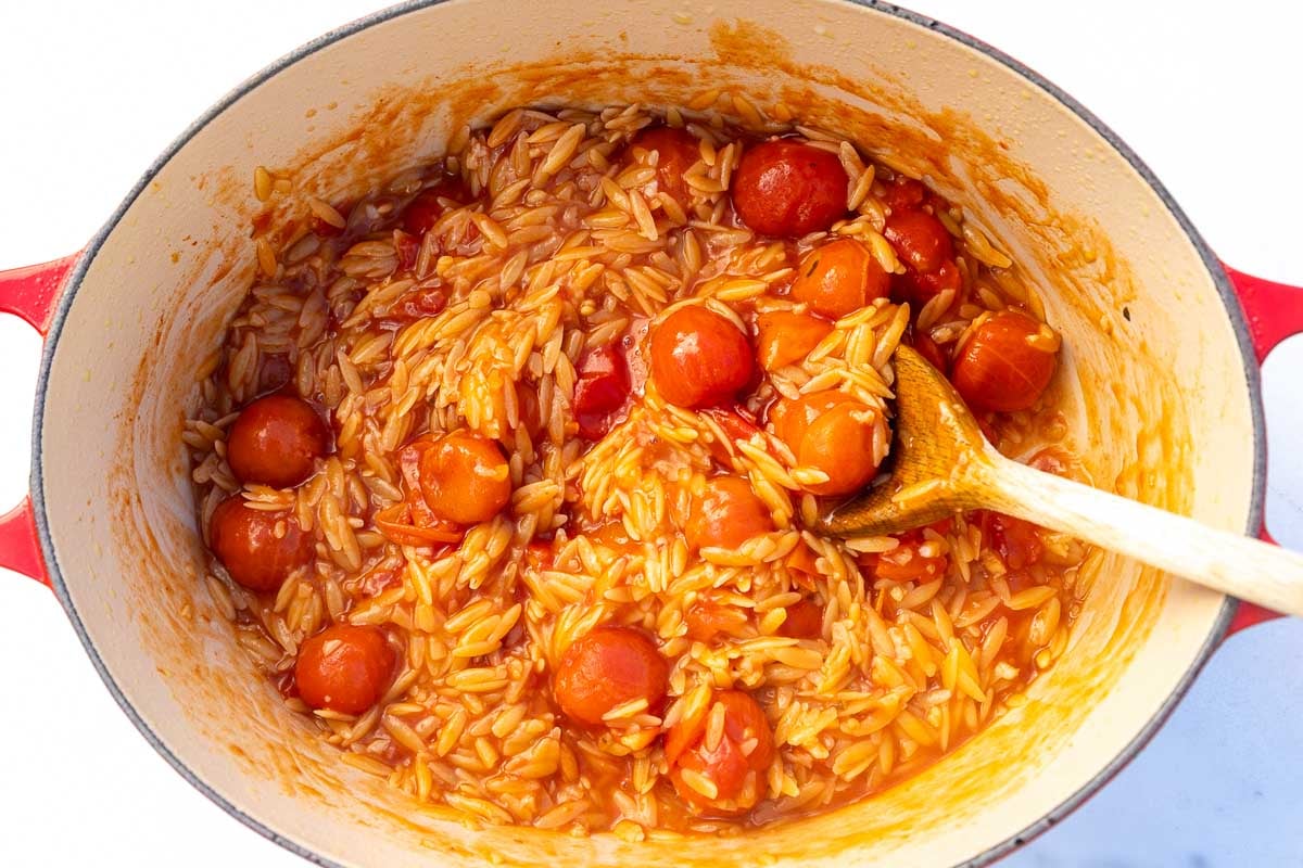 An overhead view of tomato orzo in a red cast iron pot, with a wooden spoon.