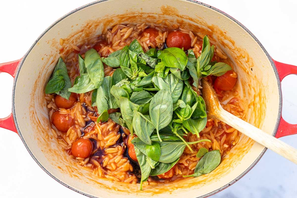 Overhead view of a red cast iron pot with tomato orzo and fresh basil in it, along with a wooden spoon.
