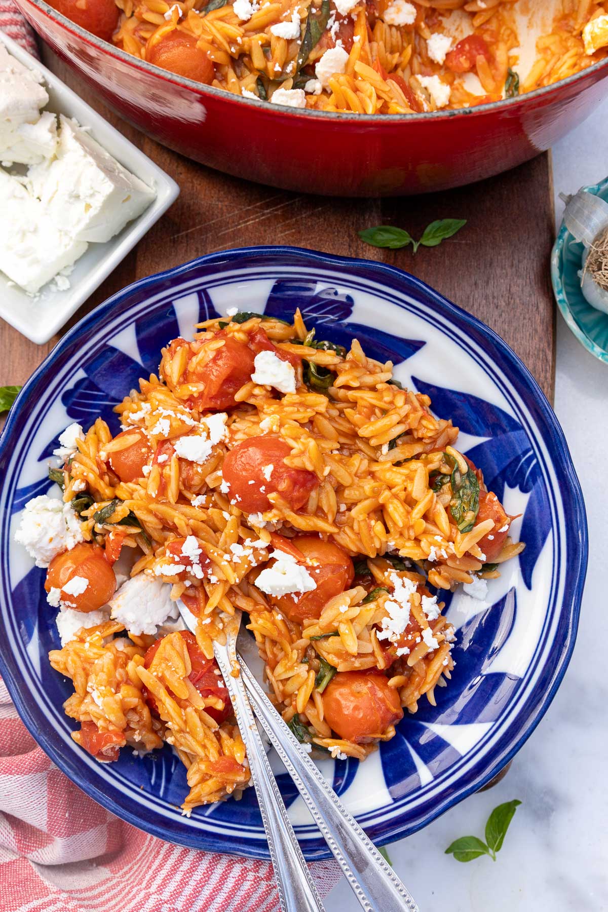 An overhead view of a blue and white bowl with a portion of tomato orzo with feta, with a knife and fork in it.