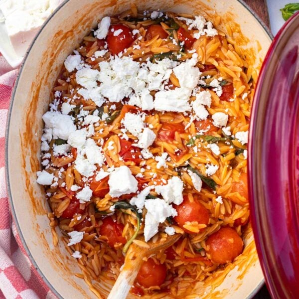 A closeup overhead of tomato orzo in a red pot with feta cheese scattered on top, the lid of the pot showing at the side and a wooden spoon.