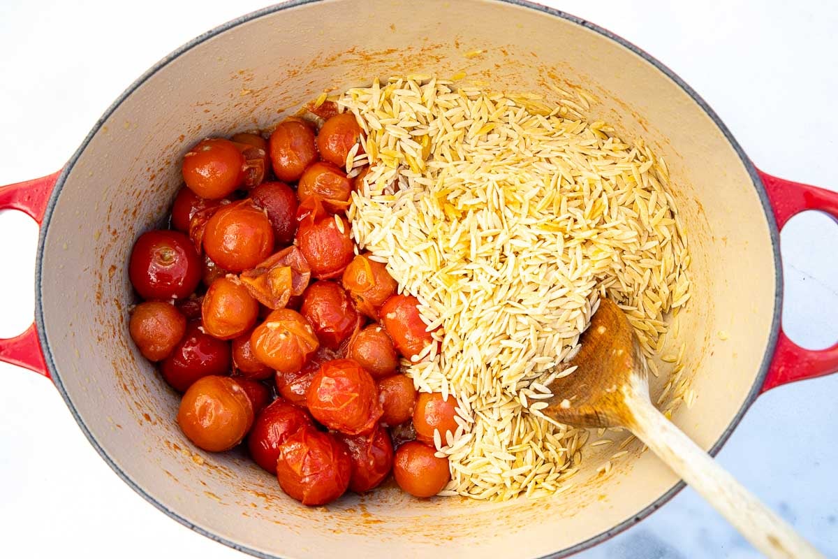 Overhead view of cooked cherry tomatoes and garlic, with dry orzo to the right, with a wooden spoon in a cast iron pot.