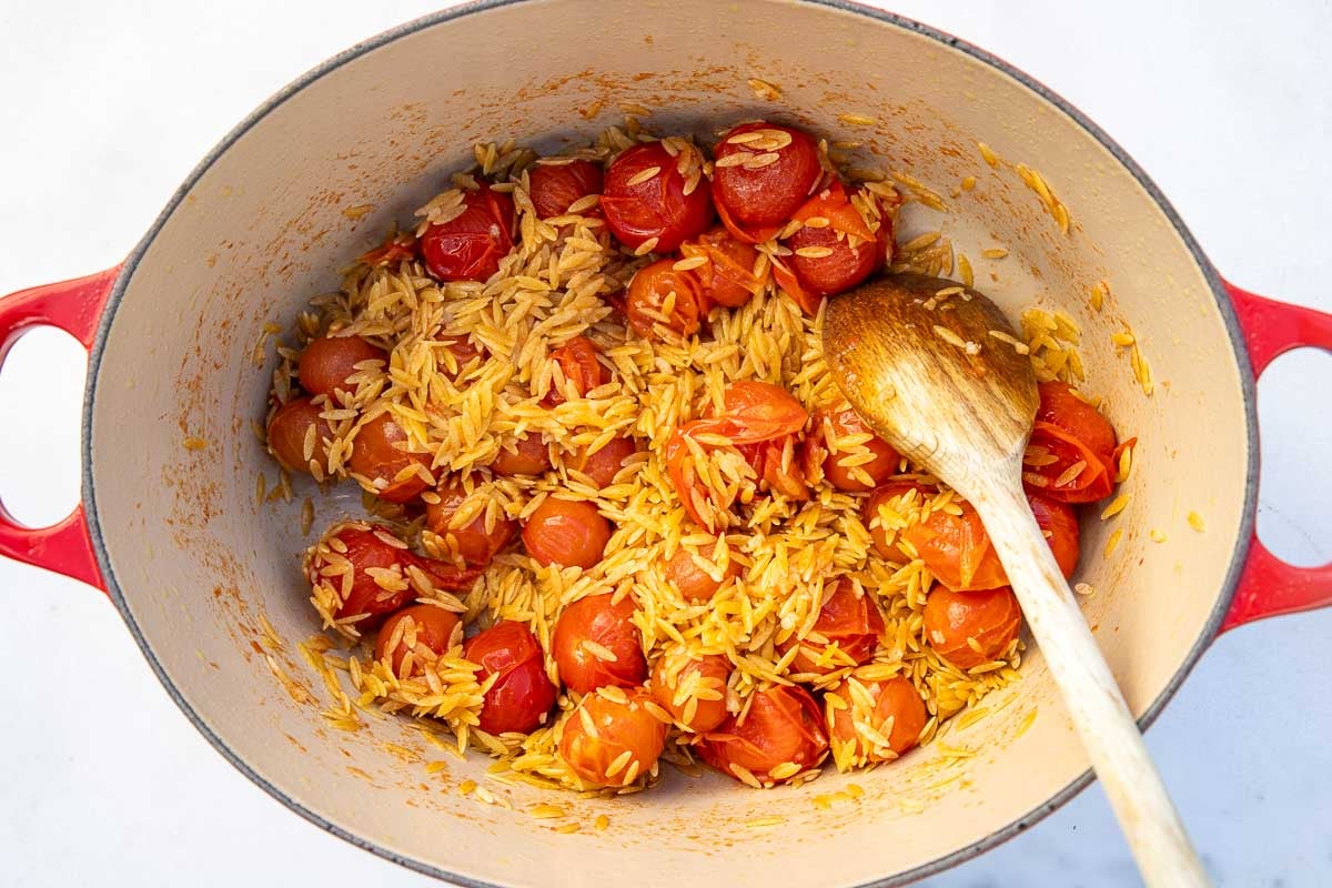 Cherry tomatoes, garlic and dry orzo mixed together in a red cast iron pot, with a wooden spoon.