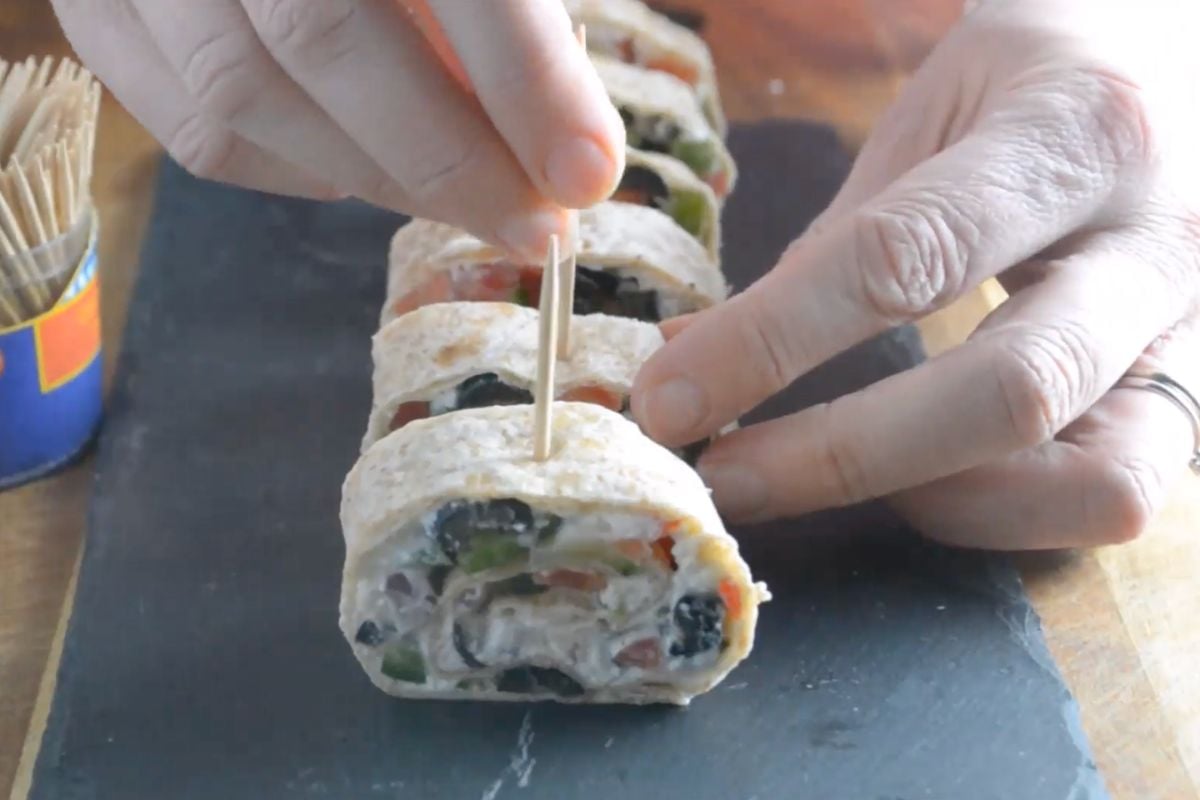 Someone putting cocktail sticks into the top of tortilla pinwheels on a black slate board.