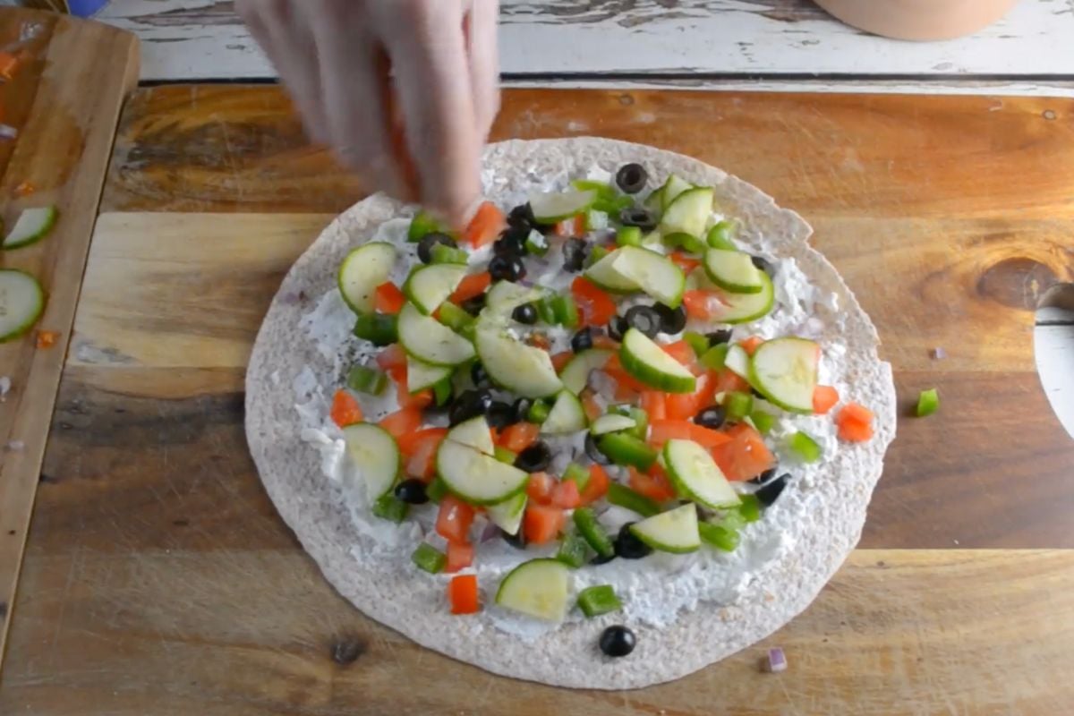 Someone sprinkling Greek salad ingredients over a tortilla spread with cream cheese.