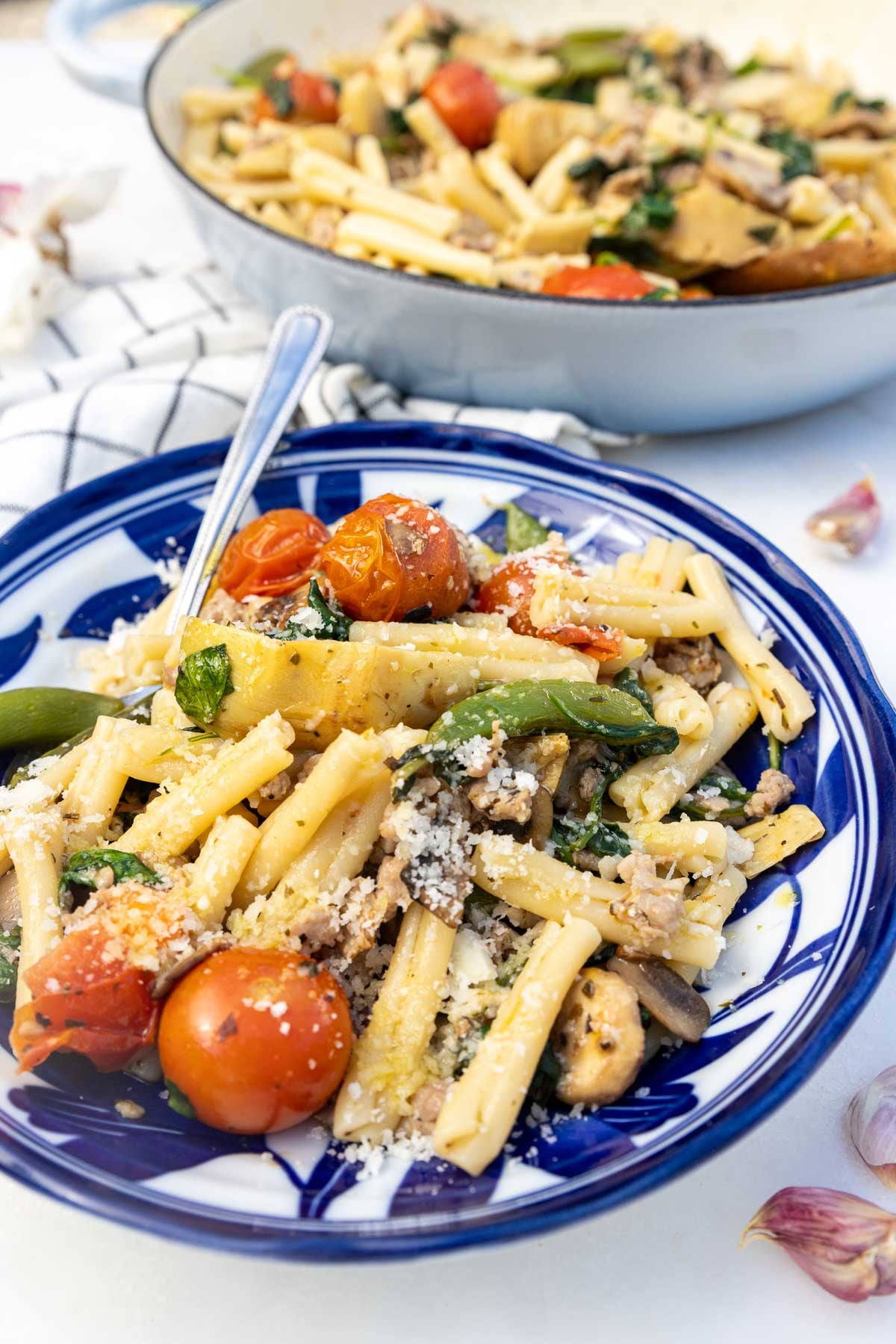 Closeup of a white and blue bowl of Italian sausage pasta with a spoon in it and the rest of the pasta in a pan behind.