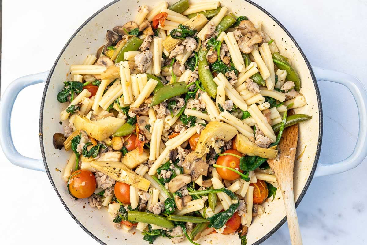 Colorful Italian sausage and vegetable pasta in a large pan with a wooden spoon.