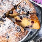 Closeup of the edge of the top of a blueberry yogurt cake with two pieces cut out and a purple tea towel above it.