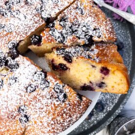 Closeup of the edge of the top of a blueberry yogurt cake with two pieces cut out and a purple tea towel above it.