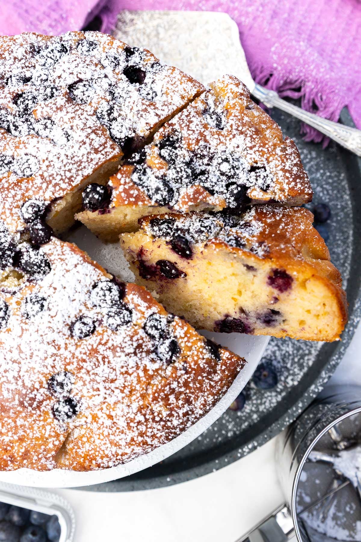 Closeup of the edge of the top of a blueberry yogurt cake with two pieces cut out and a purple tea towel above it.