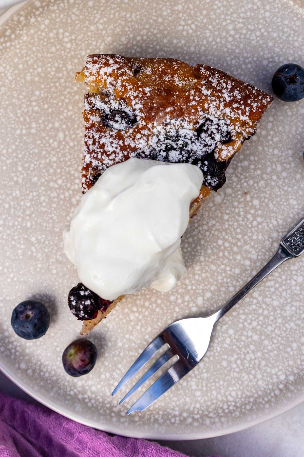 Overhead view of a piece of the cake on a neutral colored plate, with yogurt on top and blueberries and a fork at the side.