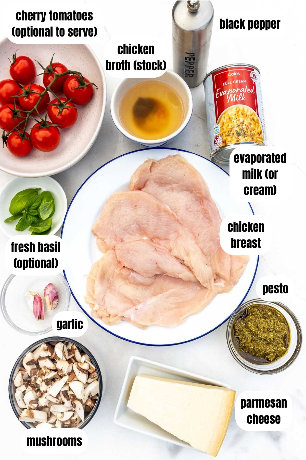 An overhead view of all the ingredients needed to make pesto chicken, including chicken breasts, mushrooms, evaporated milk, parmesan cheese, pesto, garlic, chicken broth, black pepper, cherry tomatoes to serve and optional fresh basil.